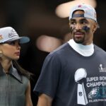 Two fans walk together, a woman in a gray vest and cap beside a man wearing a Super Bowl Champions shirt and cap, looking at each other.