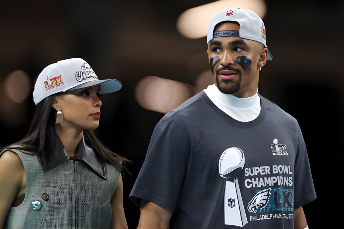 Two fans walk together, a woman in a gray vest and cap beside a man wearing a Super Bowl Champions shirt and cap, looking at each other.